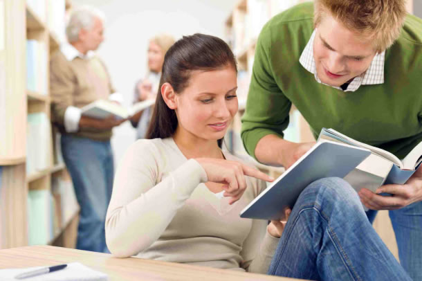 Photo of two teens in the library, one with hard cover book and another using a iPad
